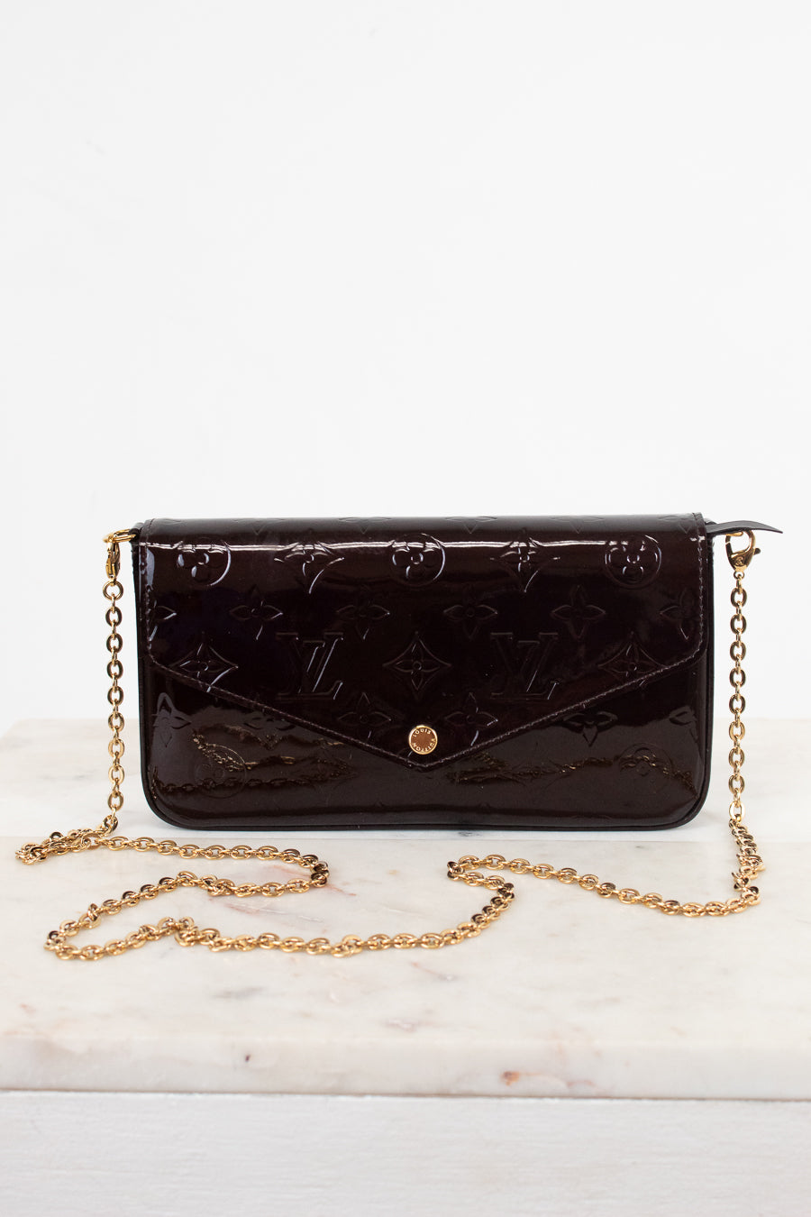 A dark burgundy patent leather clutch with a gold chain strap., isolated on a white background.