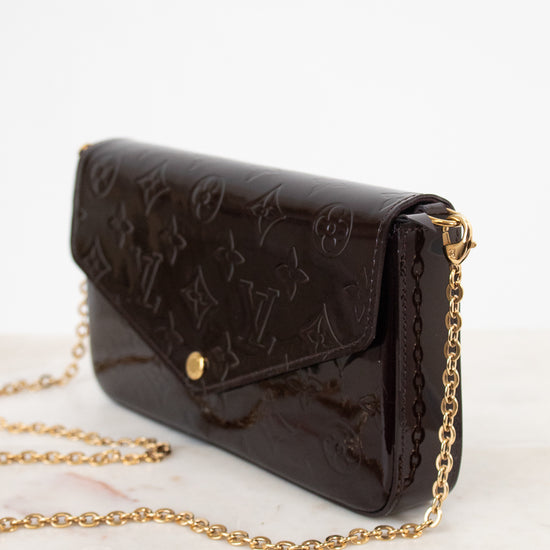 A dark brown Louis Vuitton monogram coated canvas clutch bag with a gold-tone chain strap, resting on a light marble surface against a white background.