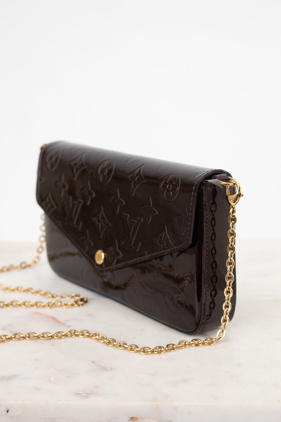 A dark brown Louis Vuitton monogram coated canvas clutch bag with a gold-tone chain strap, resting on a light marble surface against a white background.