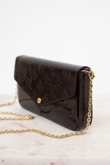 A dark brown Louis Vuitton monogram coated canvas clutch bag with a gold-tone chain strap, resting on a light marble surface against a white background.