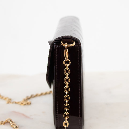 A close-up side view of a dark burgundy patent leather clutch bag with a gold chain strap, resting on a light marble surface against a white background.