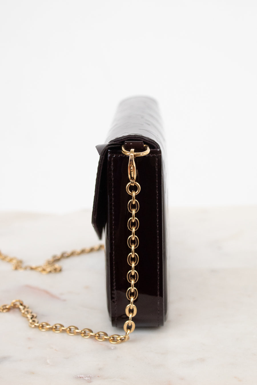 A close-up side view of a dark burgundy patent leather clutch bag with a gold chain strap, resting on a light marble surface against a white background.