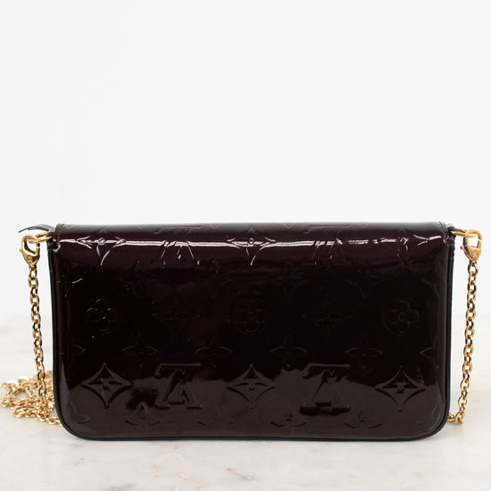 A dark burgundy Louis Vuitton patent leather clutch bag with an embossed monogram pattern and a gold chain strap, resting on a white marble surface against a white background.