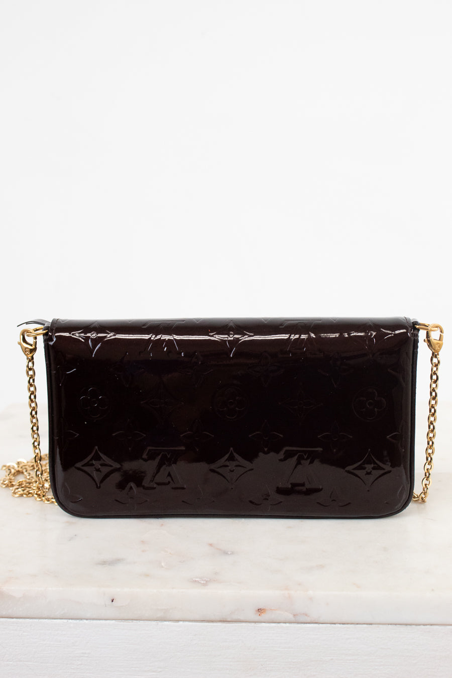 A dark burgundy Louis Vuitton patent leather clutch bag with an embossed monogram pattern and a gold chain strap, resting on a white marble surface against a white background.