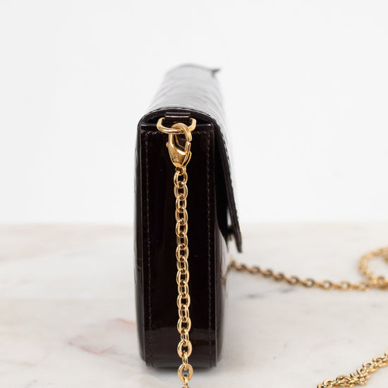 A close-up side view of a dark burgundy embossed leather clutch bag with a gold-tone chain strap, resting on a white marble surface against a white background.