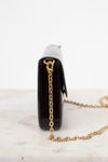 A close-up side view of a dark burgundy embossed leather clutch bag with a gold-tone chain strap, resting on a white marble surface against a white background.
