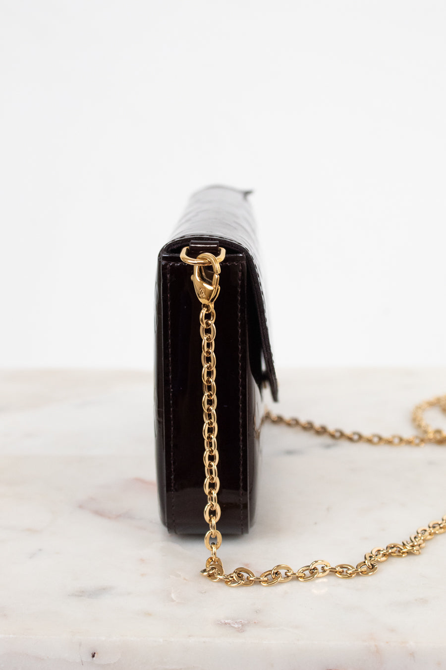 A close-up side view of a dark burgundy embossed leather clutch bag with a gold-tone chain strap, resting on a white marble surface against a white background.