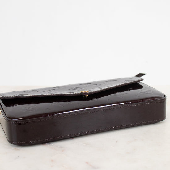 A dark burgundy glossy patent leather clutch bag with a textured flap, resting on a white marble surface against a white background.