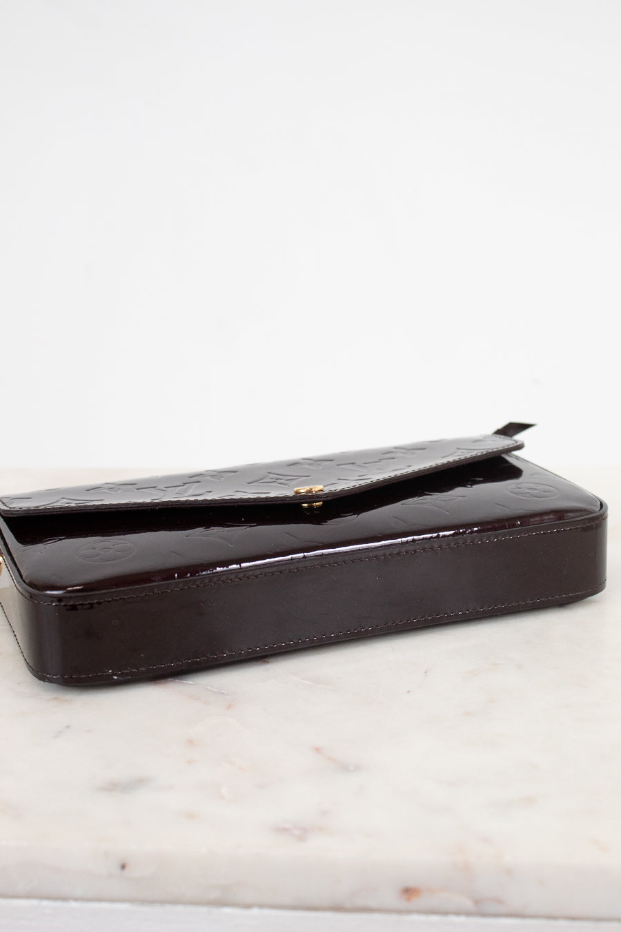 A dark burgundy glossy patent leather clutch bag with a textured flap, resting on a white marble surface against a white background.