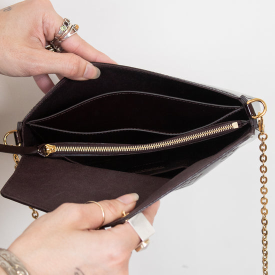A close-up of a person's hands holding open a dark burgundy textured leather clutch bag with gold-tone hardware, a zippered compartment, and a gold chain strap against a plain white background.