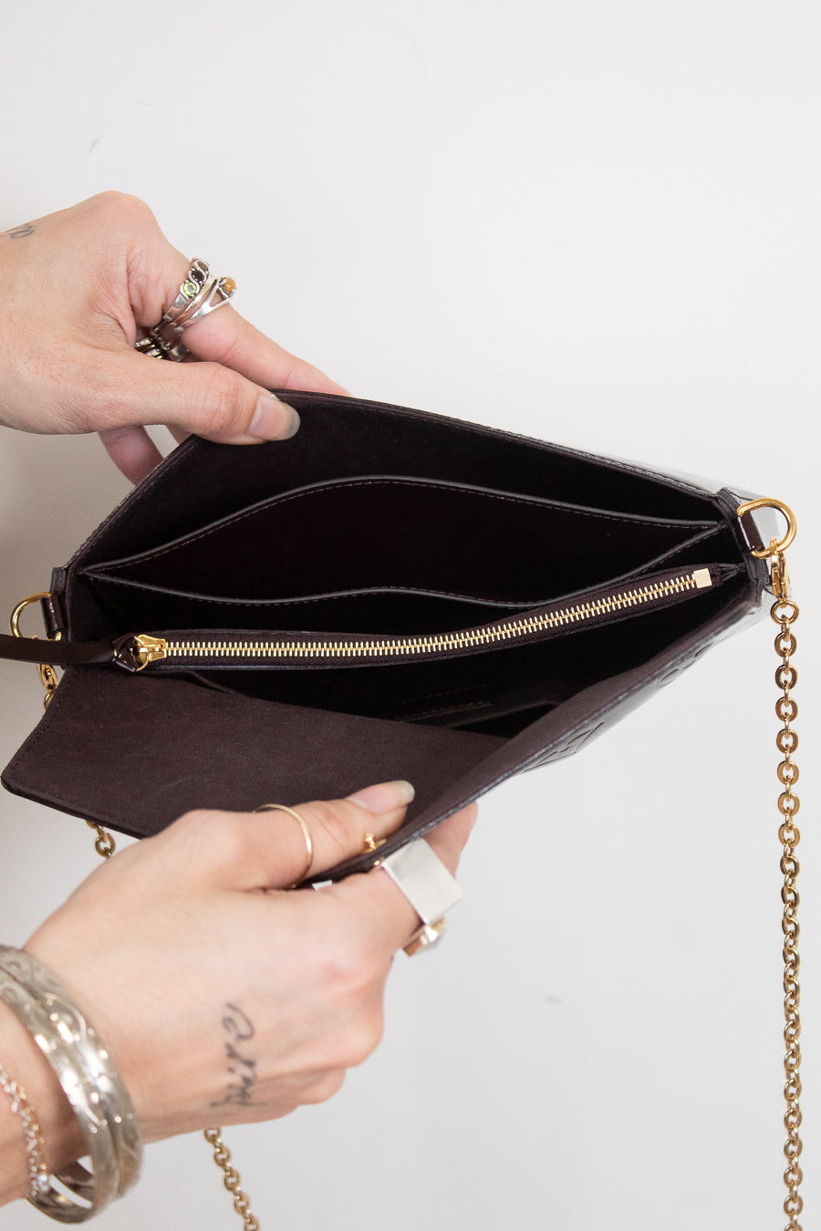 A close-up of a person's hands holding open a dark burgundy textured leather clutch bag with gold-tone hardware, a zippered compartment, and a gold chain strap against a plain white background.