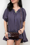 A woman wearing a dark purple puff-sleeve blouse with a ruffled hem, holding a dark burgundy patent leather clutch with a gold chain strap.