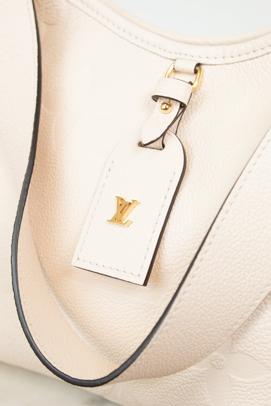 Close-up of a cream-colored leather Louis Vuitton shoulder bag with a matching leather luggage tag featuring a gold-tone LV logo, set against a light background.