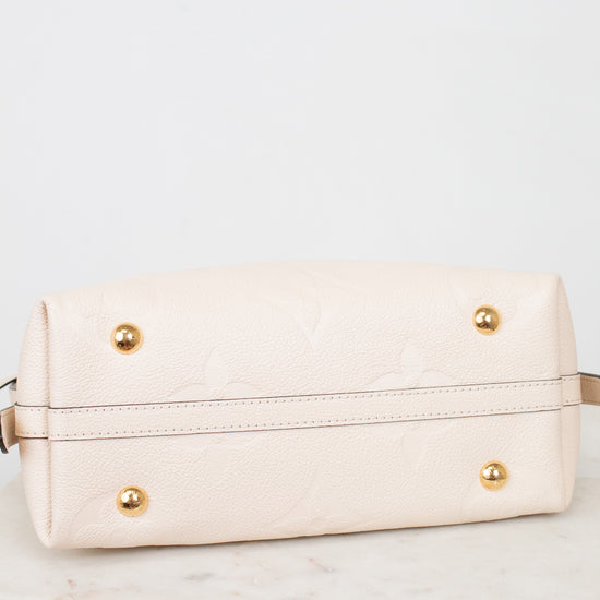 A cream-colored leather handbag shown from the bottom, featuring a subtle embossed floral pattern, gold-tone studs, and a horizontal stitched seam, resting on a white marble surface against a white background.