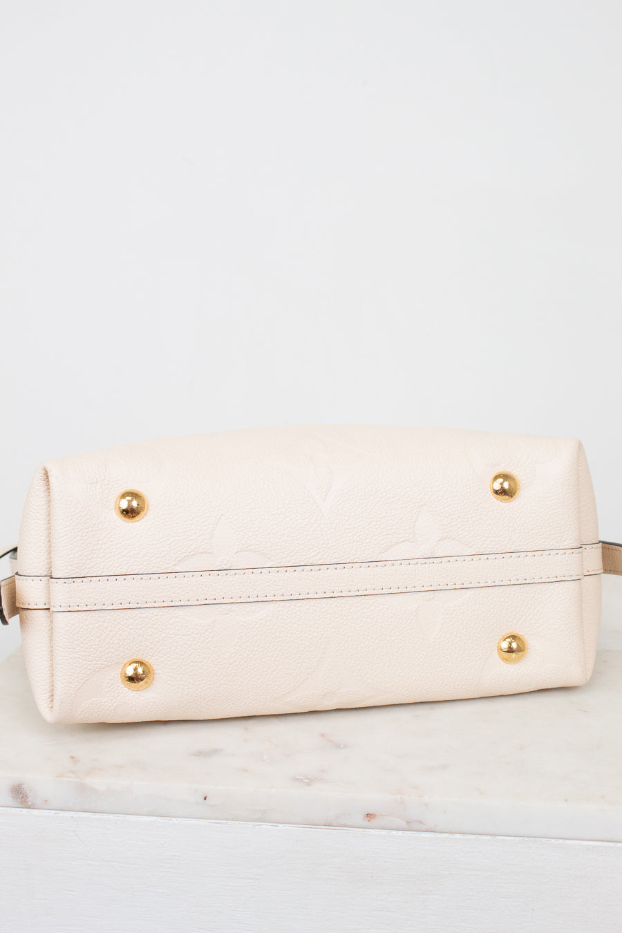 A cream-colored leather handbag shown from the bottom, featuring a subtle embossed floral pattern, gold-tone studs, and a horizontal stitched seam, resting on a white marble surface against a white background.