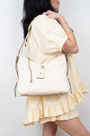 A woman wearing a yellow and white gingham dress carries a cream-colored leather shoulder bag with a small gold logo on the front, photographed against a plain white background.