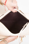 Close-up of a hand holding open a large off-white handbag to show a dark brown interior lining, with gold-tone hardware and a white strap visible against a plain light background.
