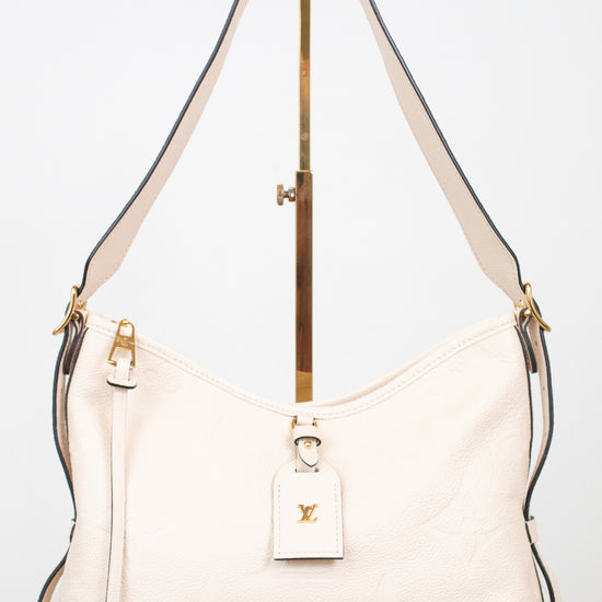 A cream-colored Louis Vuitton shoulder bag with gold-tone hardware and a matching leather hangtag, displayed on a stand against a white background.