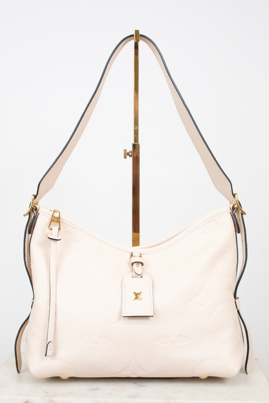 A cream-colored Louis Vuitton shoulder bag with gold-tone hardware and a matching leather hangtag, displayed on a stand against a white background.