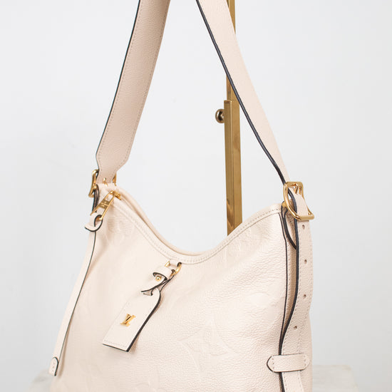 A cream-colored pebbled leather hobo shoulder bag with gold-tone hardware, displayed on a wooden stand against a light gray background.