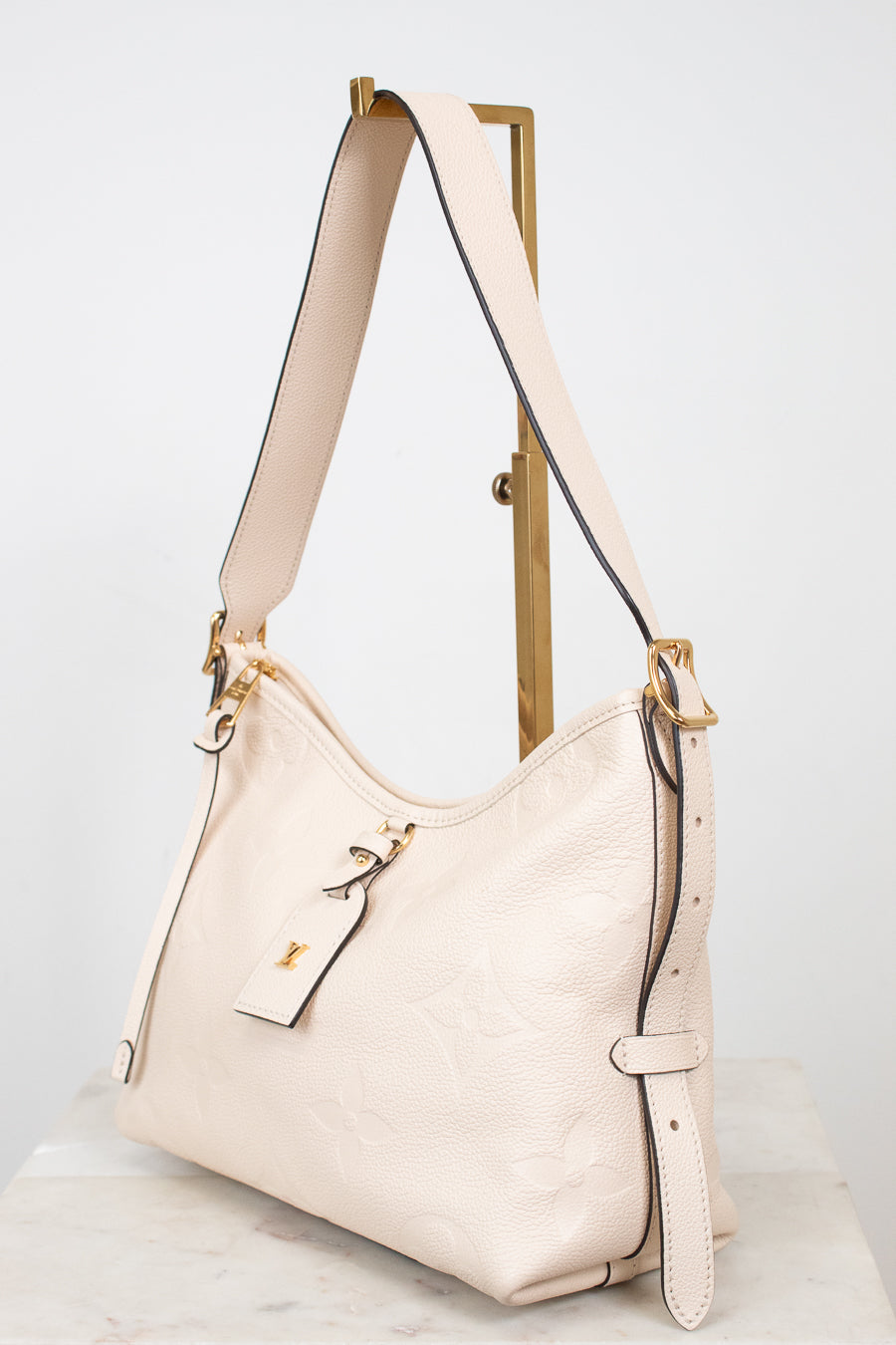 A cream-colored pebbled leather hobo shoulder bag with gold-tone hardware, displayed on a wooden stand against a light gray background.