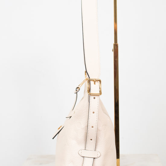 A light beige pebbled leather shoulder bag with gold-tone hardware, displayed on a white marble surface next to a gold metal stand against a white background.