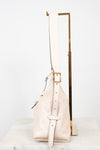 A light beige pebbled leather shoulder bag with gold-tone hardware, displayed on a white marble surface next to a gold metal stand against a white background.