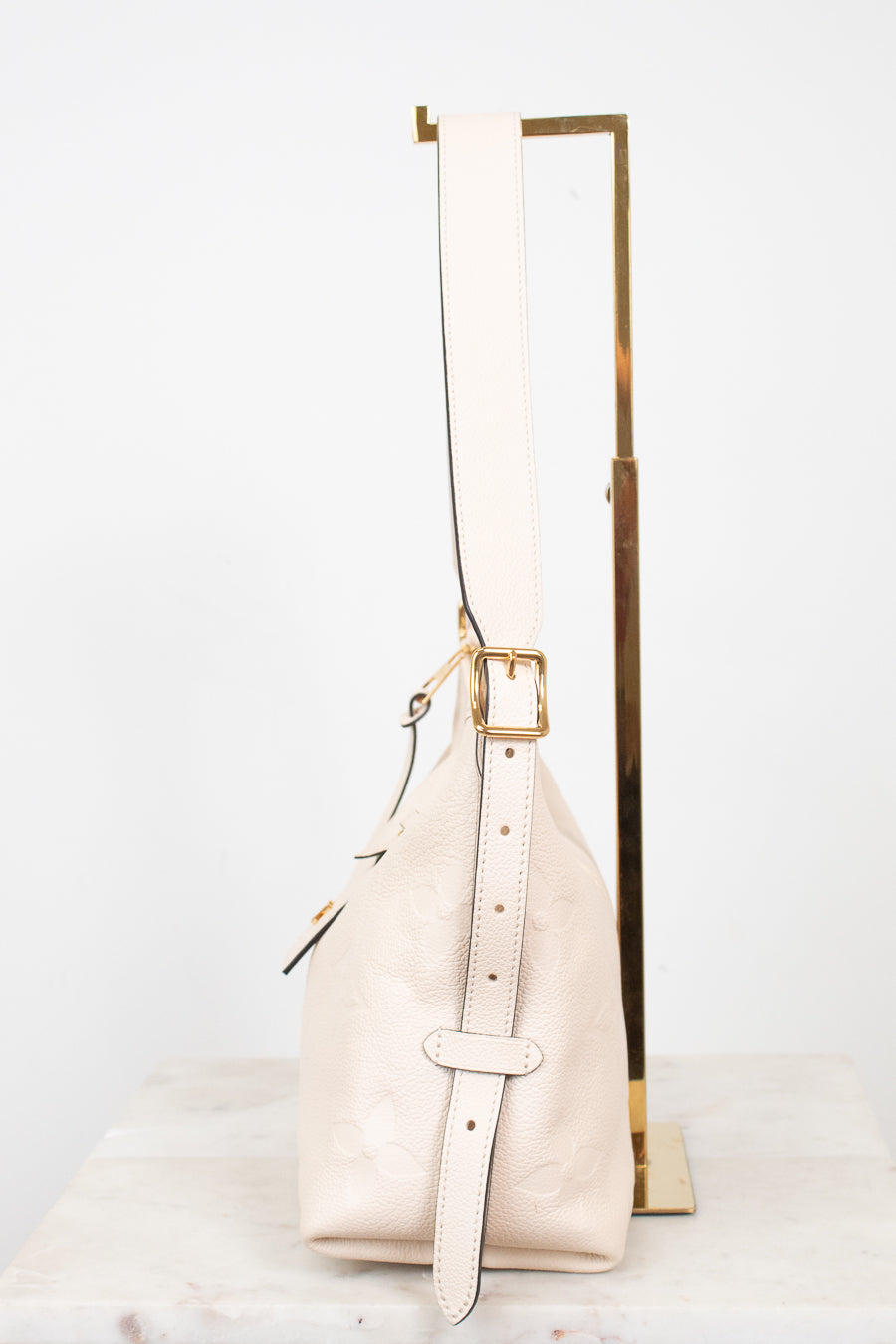 A light beige pebbled leather shoulder bag with gold-tone hardware, displayed on a white marble surface next to a gold metal stand against a white background.
