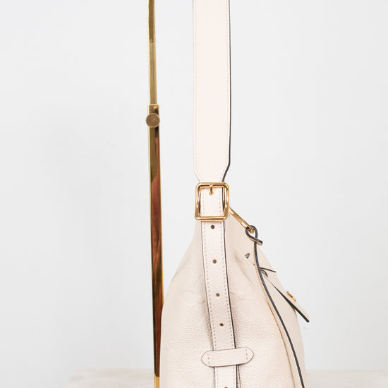 A light cream leather shoulder bag with gold-tone hardware, displayed on a minimalist gold metal stand against a clean white background.