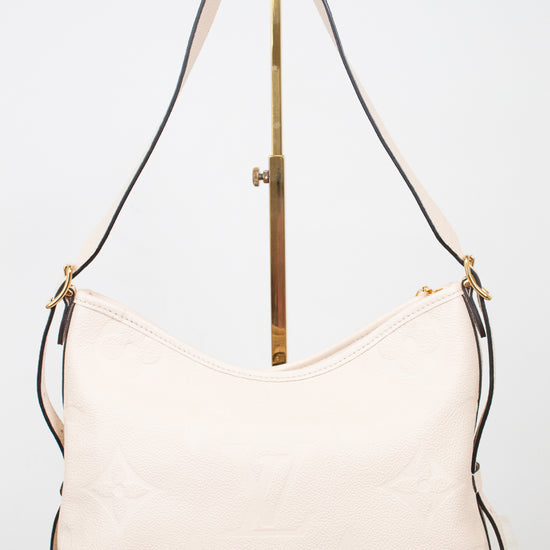 A light cream-colored leather shoulder bag with a subtle embossed monogram pattern, displayed on a gold-colored stand against a white background.