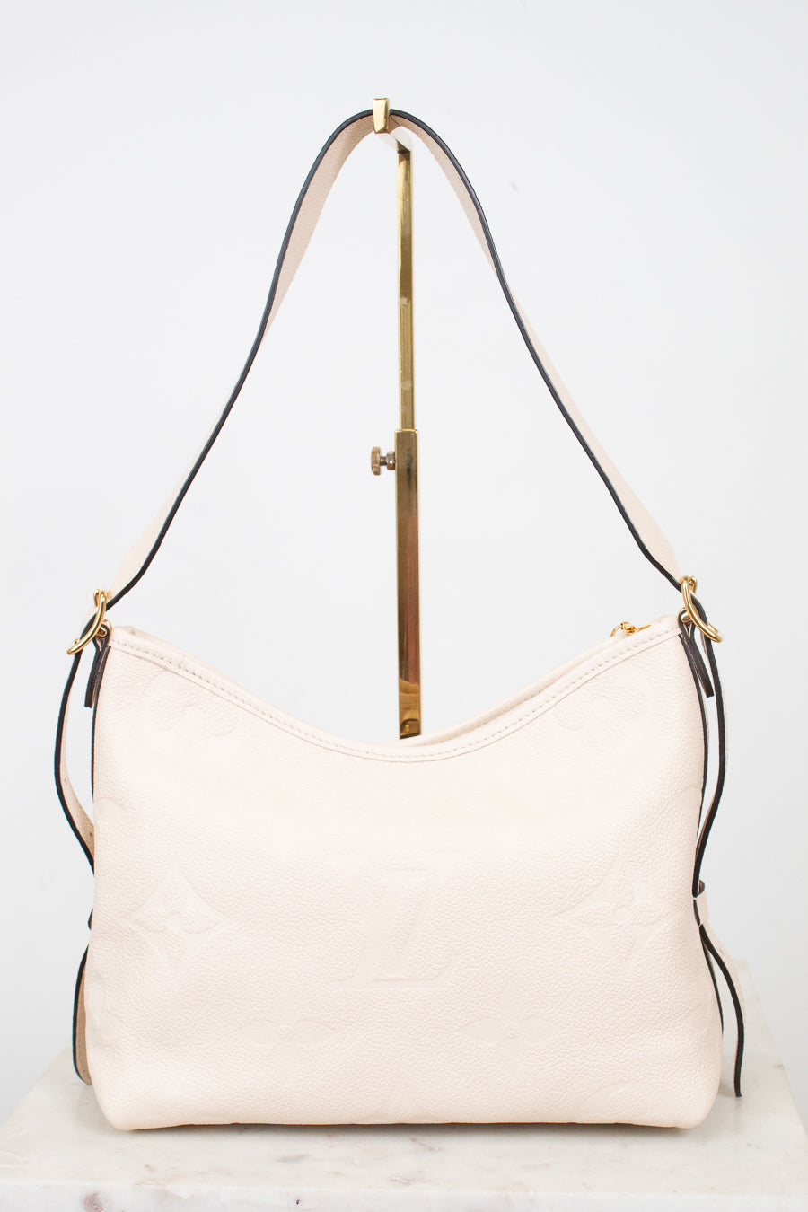 A light cream-colored leather shoulder bag with a subtle embossed monogram pattern, displayed on a gold-colored stand against a white background.