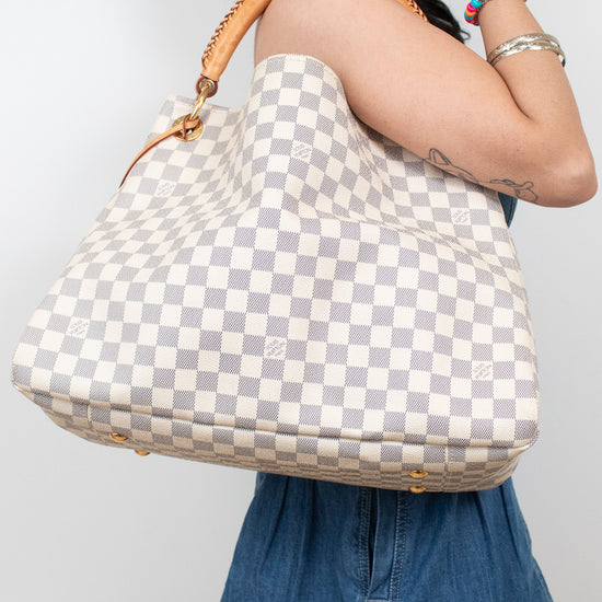 A woman wearing denim carries a large Louis Vuitton hobo bag in the brand's signature Damier Azur checkered pattern with a tan leather shoulder strap and gold-tone hardware.