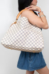 A woman wearing denim carries a large Louis Vuitton hobo bag in the brand's signature Damier Azur checkered pattern with a tan leather shoulder strap and gold-tone hardware.