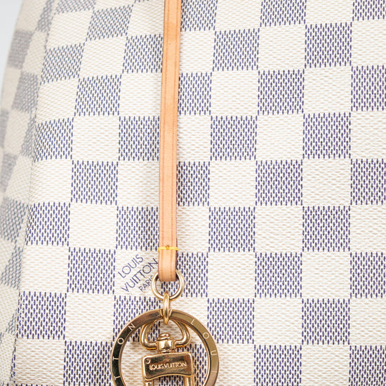 Close-up of a Louis Vuitton handbag in Damier Azur canvas, featuring a cream and blue-gray checkered pattern, a light tan leather strap, and a gold-tone interlocking O-ring charm with a small Louis Vuitton logo plaque.