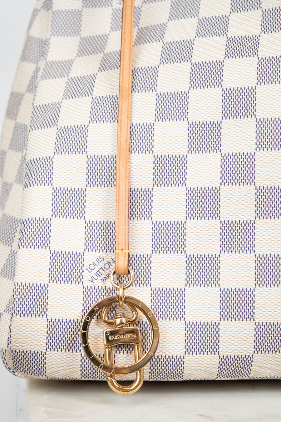 Close-up of a Louis Vuitton handbag in Damier Azur canvas, featuring a cream and blue-gray checkered pattern, a light tan leather strap, and a gold-tone interlocking O-ring charm with a small Louis Vuitton logo plaque.