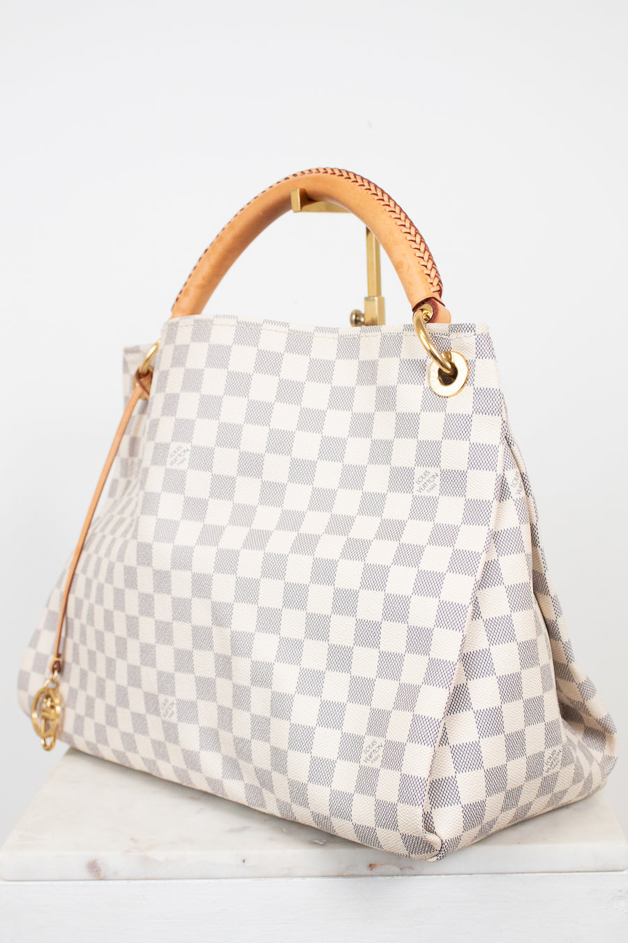 A Louis Vuitton Damier Azur canvas handbag with a light gray and white checkered pattern, tan leather top handles, and gold-tone hardware, displayed on a white marble surface against a white background.