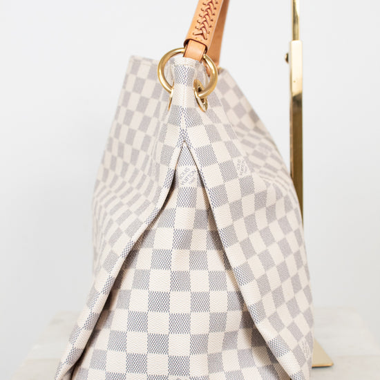 A Louis Vuitton Damier Azur canvas handbag with a beige and gray checkerboard pattern, featuring a tan leather top handle and gold-tone hardware, displayed on a stand.