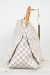 A Louis Vuitton Damier Azur canvas handbag with a beige and gray checkerboard pattern, featuring a tan leather top handle and gold-tone hardware, displayed on a stand.