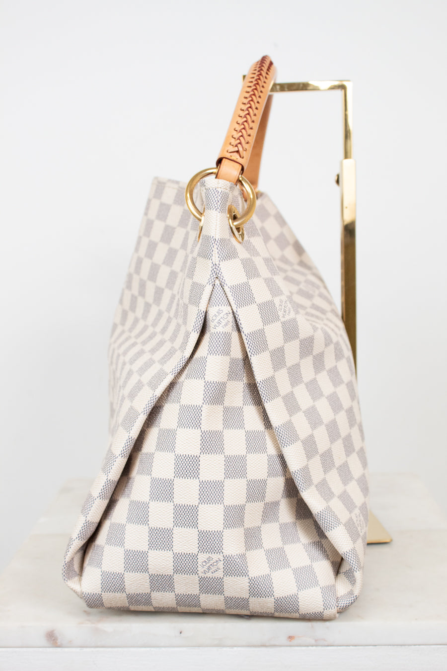 A Louis Vuitton Damier Azur canvas handbag with a beige and gray checkerboard pattern, featuring a tan leather top handle and gold-tone hardware, displayed on a stand.