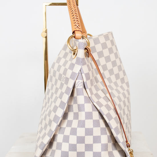 A Louis Vuitton handbag in the Damier Azur checkered canvas, featuring a light gray and off-white pattern, tan leather handle, and gold-tone hardware, displayed on a white marble surface against a white background.