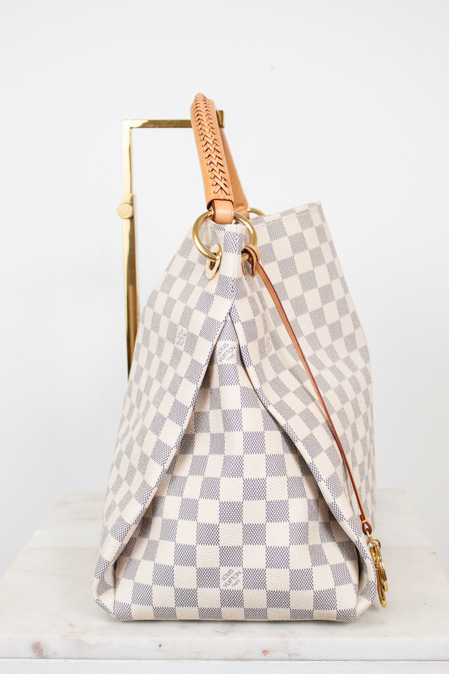 A Louis Vuitton handbag in the Damier Azur checkered canvas, featuring a light gray and off-white pattern, tan leather handle, and gold-tone hardware, displayed on a white marble surface against a white background.