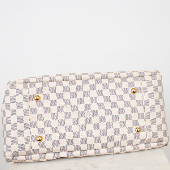 Bottom view of Louis Vuitton bag with a light gray and off-white checkerboard pattern, featuring four gold-tone metal studded feet, displayed on a white marble block against a white background.