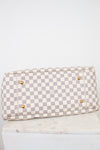 Bottom view of Louis Vuitton bag with a light gray and off-white checkerboard pattern, featuring four gold-tone metal studded feet, displayed on a white marble block against a white background.