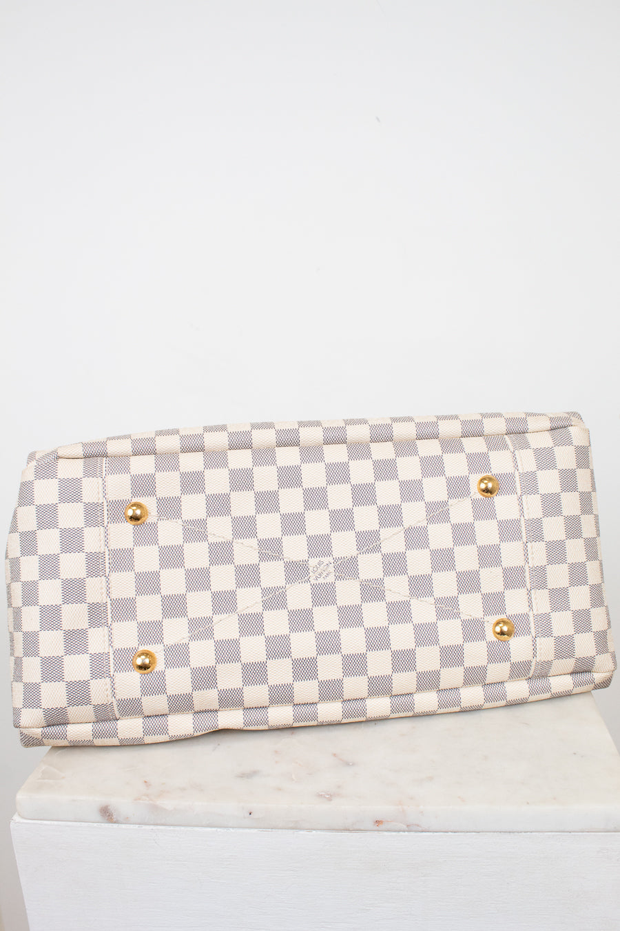 Bottom view of Louis Vuitton bag with a light gray and off-white checkerboard pattern, featuring four gold-tone metal studded feet, displayed on a white marble block against a white background.