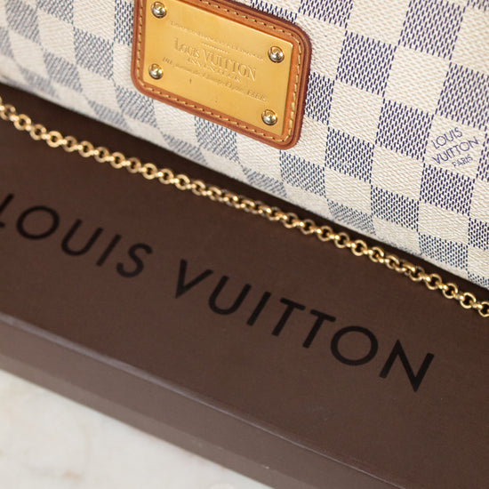 A close-up of a Louis Vuitton monogram bag featuring the Damier Azur check pattern, a gold-tone metal logo plate, and a gold chain strap, resting on a brown Louis Vuitton box.