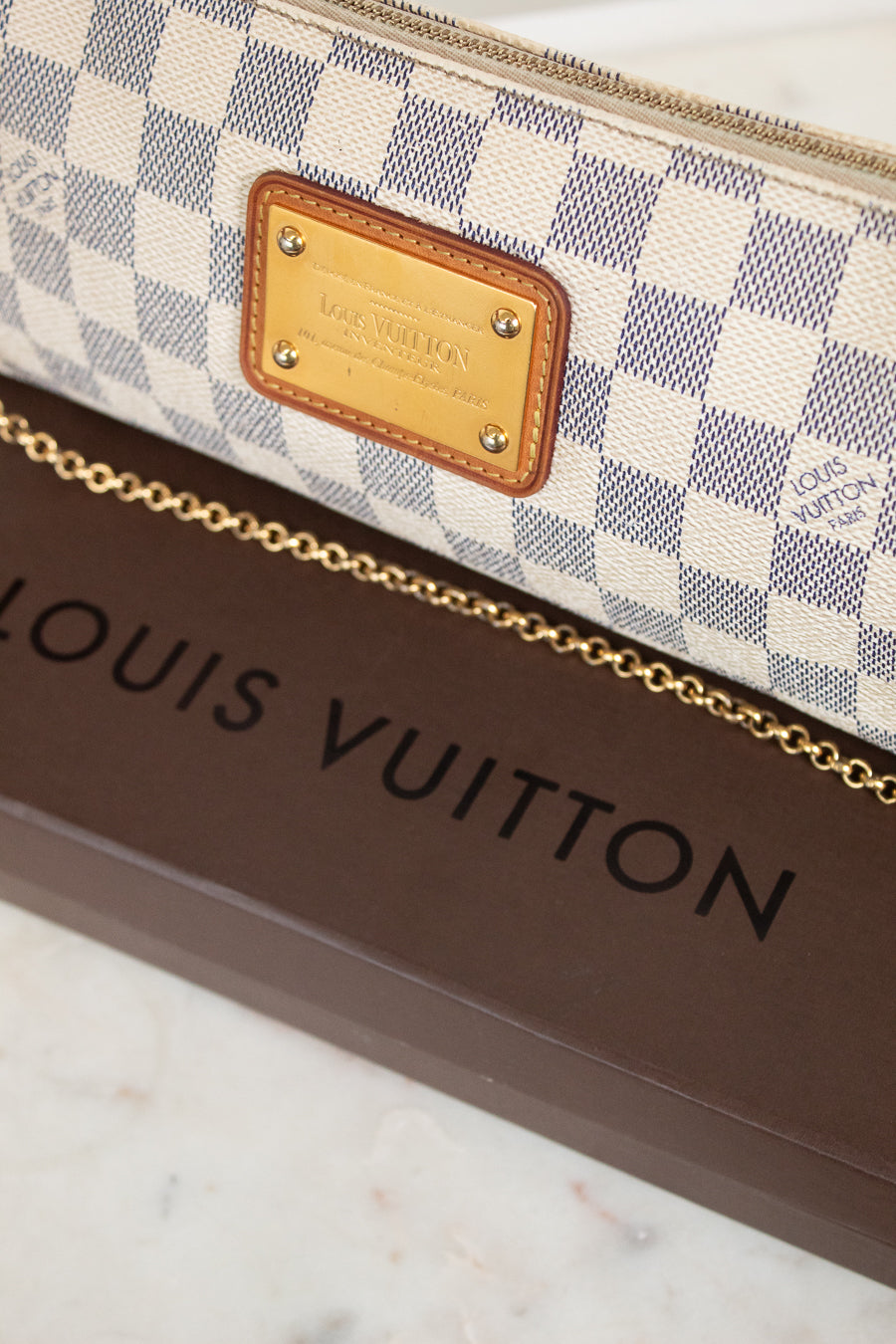A close-up of a Louis Vuitton monogram bag featuring the Damier Azur check pattern, a gold-tone metal logo plate, and a gold chain strap, resting on a brown Louis Vuitton box.