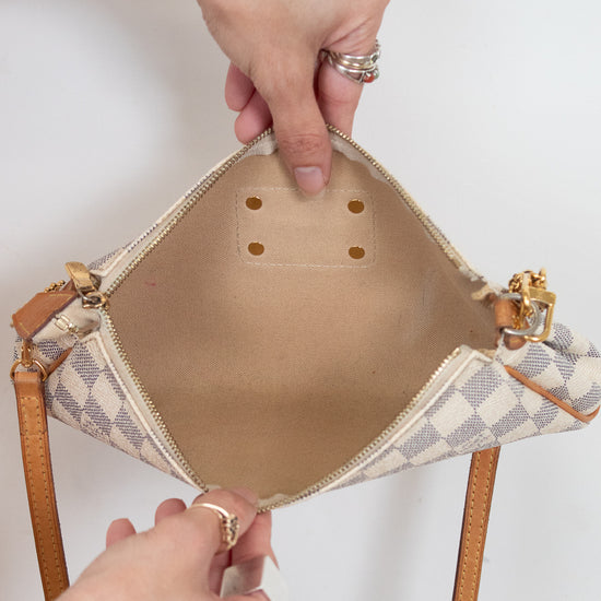 A close-up of a handbag's open interior, showing a beige and white checkered patterned exterior and beige fabric interior, held open by two hands wearing rings against a plain white background.