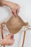 A close-up of a handbag's open interior, showing a beige and white checkered patterned exterior and beige fabric interior, held open by two hands wearing rings against a plain white background.