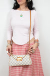 A woman wearing a light pink long-sleeve top and a red-and-white gingham skirt, holding a beige and white checkered shoulder bag with a tan strap and gold-tone hardware.