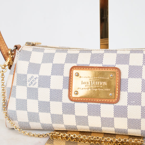 A Louis Vuitton shoulder bag in Damier Azur canvas with a blue and white checkered pattern, tan leather trim, and a gold-tone metal plaque and on a light marble background.
